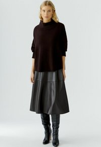 Dunkel gestrickter Pullover mit Kapuze, kombiniert mit einem schwarzen Leder-Midi-Rock und eng anliegenden schwarzen kniehohen Stiefeln. Glatte Texturen, moderne Silhouette.