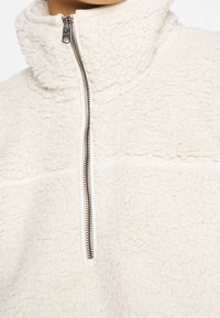 Kermanvärinen fleece-takki, jossa on hopeanvärinen vetoketju, korkea kaulus ja pehmeä, teksturoitu kangas, kuvattuna kaulasta rinnan alueelle.