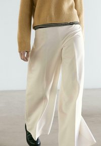 Kremowe spodnie o szerokich nogawkach o gładkiej fakturze, zestawione z beżowym, prążkowanym swetrem z czarnym paskiem na dole. Widoczne czarne obuwie.