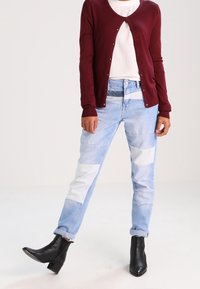 Cardigan bordeaux, t-shirt bianco, jeans patchwork blu chiaro, stivali neri alla caviglia. Il cardigan è dotato di bottoni; i jeans hanno diverse tonalità e trame.
