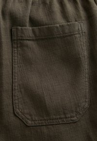 Primer plano de tela marrón que presenta un bolsillo cuadrado con costuras reforzadas y una superficie texturizada, destacando sutiles variaciones en el color.