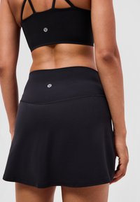 Donna che indossa un crop top sportivo nero e una gonna nera svasata con piccoli loghi bianchi sulla schiena su uno sfondo semplice.