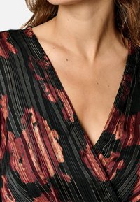 Blouse noire avec un motif floral rouge, plis verticaux et accents métalliques. Décolleté en V profond. Tissu texturé avec une finition brillante.