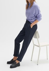 Žena op�řená o bílou židli, oblečená v svetru levandulové barvy, tmavě modrých kalhotách s manžetami a černých mokasínách na bílém pozadí.