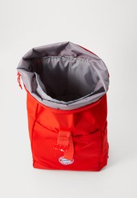 Rød nylon ryggsekk med grå foring, rull-topp lukking, frontlomme og logo lapp. Glatt tekstur og slitesterk konstruksjon.