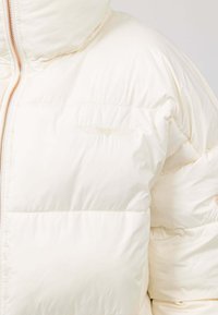 Dikke crèmekleurige jas met capuchon, met een ritssluiting en een subtiele geborduurde design op de borst in een lichte, reflecterende draad.