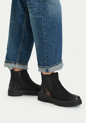 Black ankle boots with side zipper and brown detail worn with rolled-up blue denim jeans on light background.