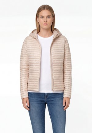 Donna che indossa una giacca imbottita con cappuccio color beige chiaro sopra una camicia bianca e jeans blu, in piedi davanti a uno sfondo bianco.