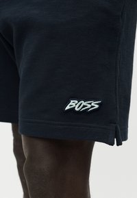 Σκούρο δέρμα πόδι που φοράει μαύρα σορτς με λευκό λογότυπο "BOSS" κοντά στο στρίφωμα και ένα μικρό πλαϊνό σκίσιμο.