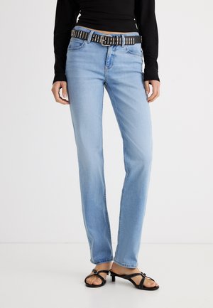 Γυναίκα που φοράει ανοιχτό μπλε τζιν παντελόνι με ίσια γραμμή, μαύρο τοπ με μακριά μανίκια, μαύρη ζώνη με καρφιά και μαύρα σανδάλια με χαμηλό τακούνι, στέκεται σε λευκό δάπεδο.