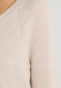 Pull en tricot beige clair à coupe ample, présentant un motif texturé et un col en V. Tissu doux avec un tissage respirant.
