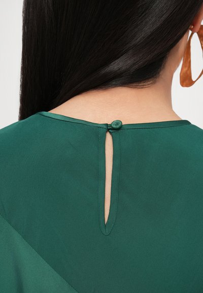 Zelená halenka s kulatým výstřihem, detailem na zádech ve formě klíčku a zeleným zapínáním na knoflíky. Měkká látka s hladkou texturou.