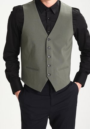 Chaleco verde oliva hecho de una tela suave, con un diseño ajustado, seis botones y dos bolsillos laterales, combinado con una camisa negra.