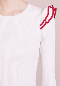 Weißes geripptes Oberteil mit strukturiertem Stoff, langen Ärmeln und einem roten rüschendetails an der Schulter mit weißen Streifen.