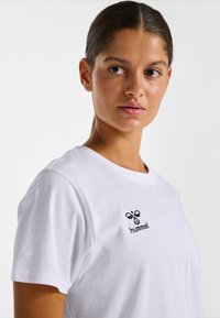 Femme aux cheveux foncés attachés, portant un t-shirt blanc Hummel, regardant légèrement vers sa gauche sur un fond uni clair.