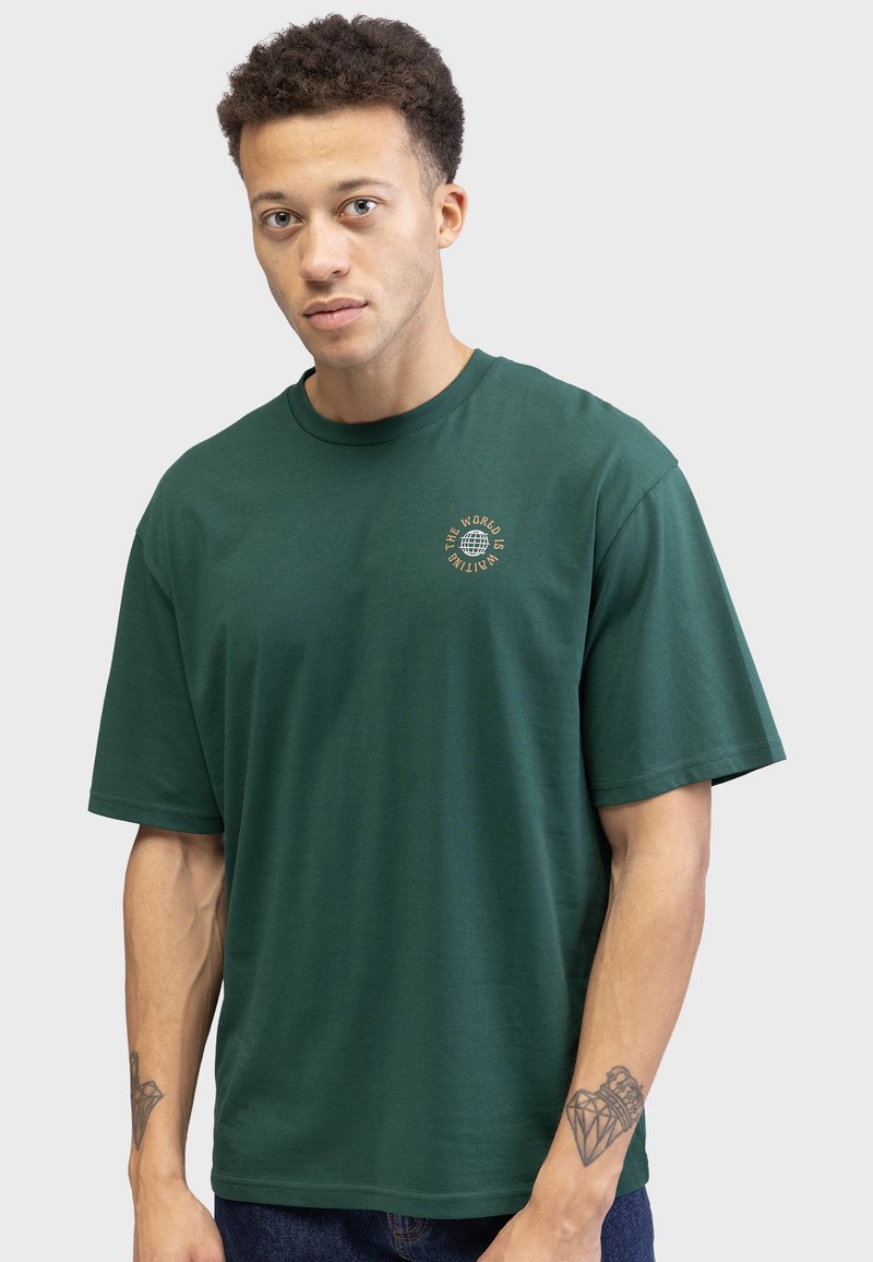 Grünes Baumwoll-T-Shirt mit Rundhalsausschnitt, kurzen Ärmeln und einem kleinen goldenen Grafiklogo auf der linken Brust. Weiche Textur, lockere Passform.