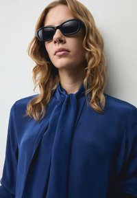Occhiali da sole neri, blusa blu navy con collo annodato, texture liscia e design fluido. Capelli castano chiaro ondulati parzialmente visibili.