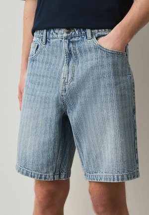 Mann trägt hellblaue, gestreifte Denim-Shorts mit Fronttaschen und einem Metallknopf, Hand in der rechten Tasche, kombiniert mit einem dunklen Hemd.