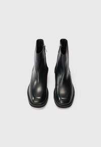 Une paire de bottes noires en cuir à chevilles avec des zips sur les côtés, des bouts carrés et des talons bas, présentée sur un fond blanc.