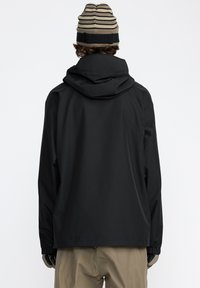 Svart vattentät jacka med huva, elastiska manschetter och en minimalistisk design. Bärs med beige byxor och en randig keps i beige och brun.