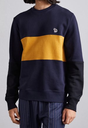 Man wearing navy sweatshirt with mustard yellow chest stripe and small multicolored elephant patch on left side.
