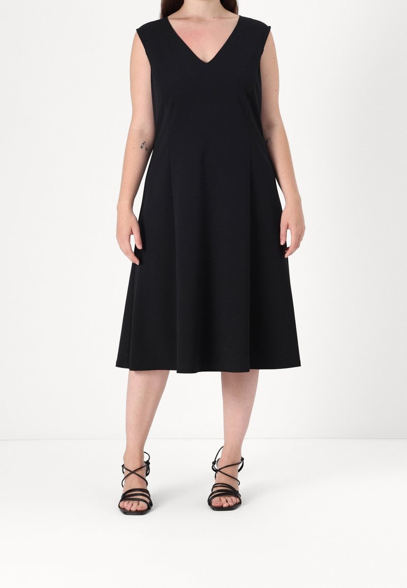 Černé bezramínkové šaty s výstřihem do V a rozšířenou sukní, vyrobené z měkké tkaniny. Doplněné černými sandály na pásku.