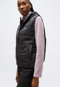 Γυναίκα που φοράει μαύρο γιλέκο χωρίς μανίκια με ραμμένο σχέδιο πάνω από ένα υφασμάτινο μωβ μακρυμάνικο τοπ, στέκεται μπροστά σε μια απλή ανοιχτή φωτεινή επιφάνεια.