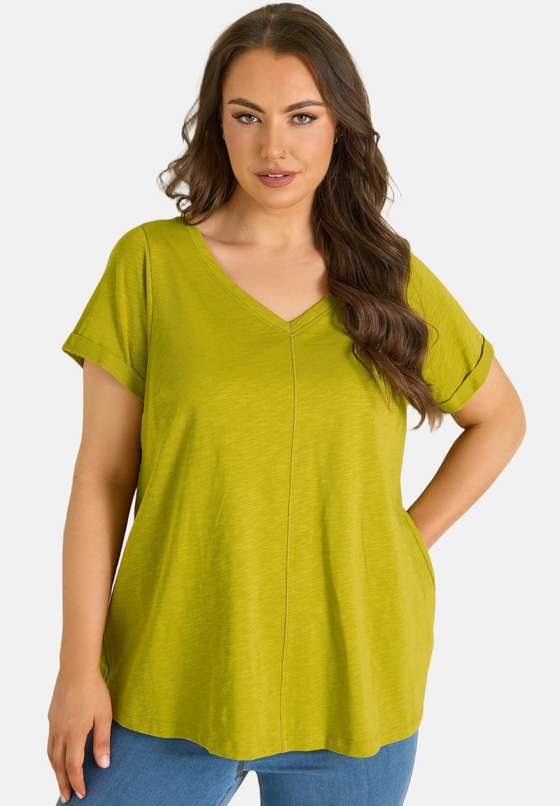 Donna con capelli castano lunghi che indossa una maglietta verde lime a maniche corte con scollo a V e dal taglio ampio, e jeans blu, in piedi con una mano sui fianchi.
