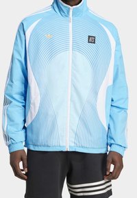 Ljusblå jacka med vita böjda linjer, hel dragkedja, hög krage och elastiska ärmslut. Har en liten svart NTS-logotyp och guldaccent.