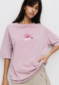 Lyserød oversized t-shirt med rund halsudskæring, der har et solgrafik og teksten "IBIZA '91" i kontrasterende farver. Blødt bomuldsstof.