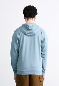 Sudadera azul claro con capucha, puños acanalados y dobladillo. Material suave, corte relajado, vista desde la espalda, sin logotipos ni patrones visibles.