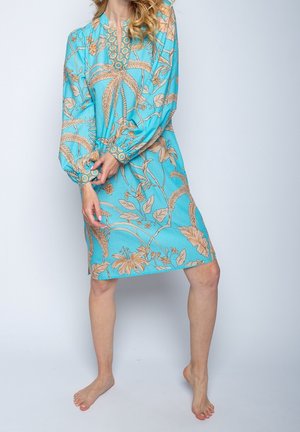 Femme portant une robe turquoise avec un motif floral et de palmiers beige, debout pieds nus sur un fond clair uni.
