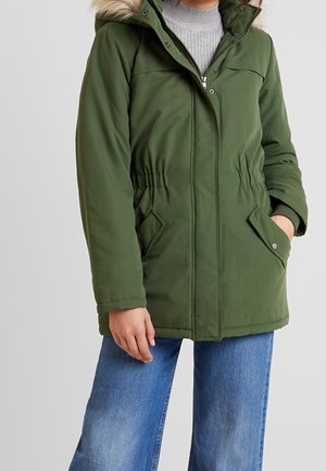 Veste d'hiver verte à capuche avec bordure en fourrure, taille élastique, poches avant, portée sur un col roulé gris et un jean bleu, avec une main dans la poche.