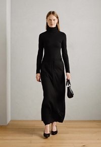 Black long-sleeve turtleneck top paired with a floor-length black skirt. The model holds a small black handbag and wears black shoes.