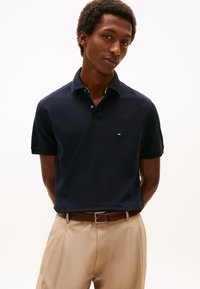 Polo azul-marinho com gola clássica e mangas curtas; apresenta logótipo pequeno no peito. Usado com calças bege claro e cinto castanho.