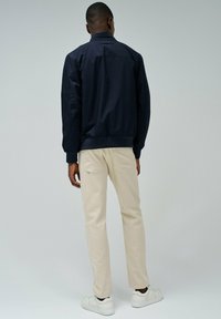 Giacca bomber navy con polsini a costine, abbinata a pantaloni beige a gamba dritta e sneakers bianche. Design minimalista con linee pulite.