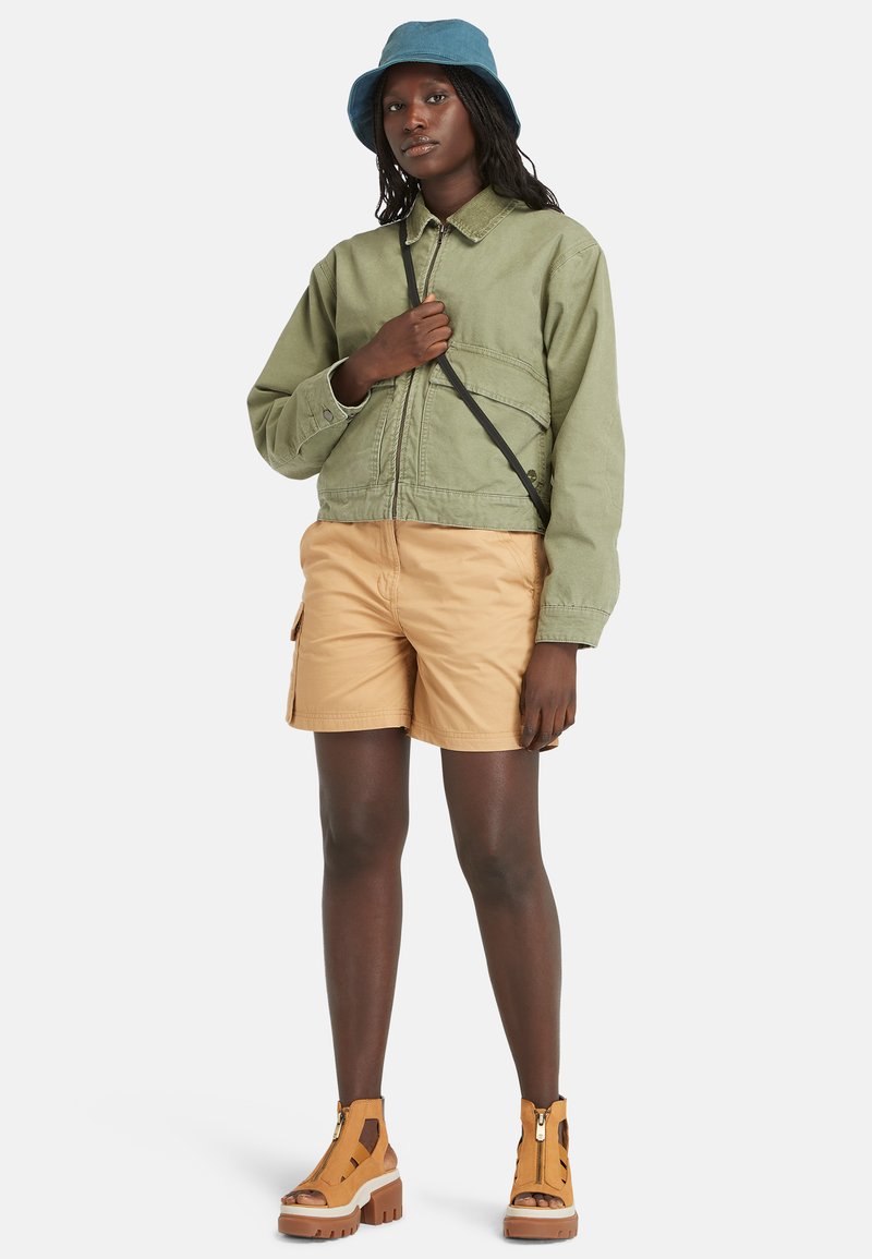 Giacca verde corta con tasche sul petto, pantaloni corti cargo beige, sandali con tacco marroni e un cappello a secchiello blu. Il look è stratificato e casual.