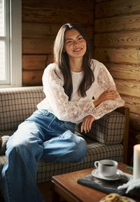 Haut blanc à manches en dentelle, jean large bleu, et collier de perles, assise sur un canapé à carreaux avec une table en bois et une tasse de café.