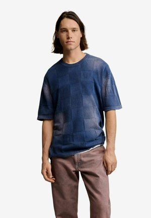 Młody mężczyzna o włosach długości do ramion, ubrany w niebieską kratkowaną koszulę z krótkim rękawem i jasnobrązowe spodnie, stojący na białym tle.