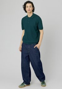 Polo shirt tricoté vert foncé à manches courtes, associé à un jean en denim bleu foncé à coupe ample. Le modèle porte des baskets vertes avec des accents blancs.