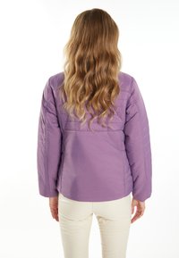 Giacca imbottita viola con una texture liscia, caratterizzata da una vestibilità morbida e cuciture orizzontali. Indossata sopra pantaloni color crema.