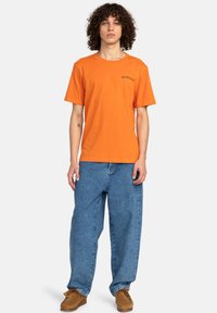 Camiseta de algodón naranja de manga corta y cuello redondo. Combinada con vaqueros de denim azul holgados y zapatos de ante marrones.