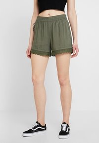 Olivově zelené šortky s elastickým pasem a krajkovou ozdobou na lemu. Lehká látka s volným střihem, doplněná černými teniskami.