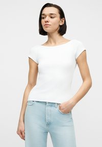 Νεαρή γυναίκα με κοντά σκούρα μαλλιά φορώντας εφαρμοστό λευκό κοντομάνικο μπλουζάκι και ανοιχτόχρωμα μπλε τζιν, με το χέρι στην τσέπη, κοιτώντας ελαφρώς στο πλάι.