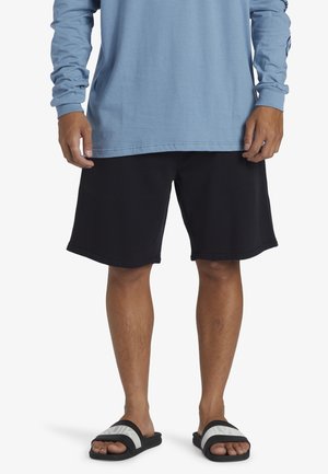 Schwarze Shorts mit geradem Schnitt und weichem Material, kombiniert mit einem hellblauen Langarmshirt und schwarzen Slidesandalen mit weißen Streifen.