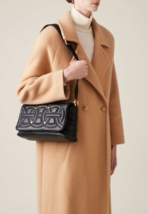 Black leather handbag with a quilted design and gold accents, held in hand. The person is wearing a beige coat and white turtleneck.