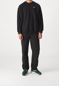 Schwarzer Sweatshirt mit runder Ausschnitt und weißem Logo, kombiniert mit dunklen Hosen und grünen Sportschuhen mit weißen Akzenten.