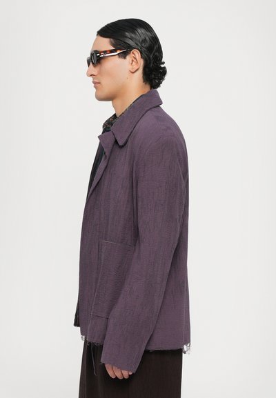 Lila strukturierte Jacke mit Kragen, Seitentaschen und fransigem Saum, getragen über einem dunkel gemusterten Hemd. Das Model trägt runde Sonnenbrillen.