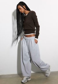 Brauner Cardigan mit langen Ärmeln über einem weißen Hemd, kombiniert mit lockeren grauen Jogginghosen. Silberne Sportschuhe runden das Outfit ab.