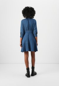 Denim dress with three-quarter sleeves, cinched waist, flared skirt, and raw hem. Worn with black ankle boots. Back view.